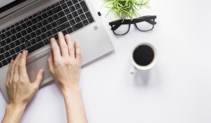 close-up-person-s-hand-typing-laptop-with-coffee-cup-eyeglasses-pot-plant-white-desk_23-2148041861