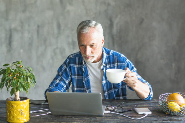 close-up-senior-man-holding-coffee-cup-using-laptop-charged-with-power-bank_23-2147859784