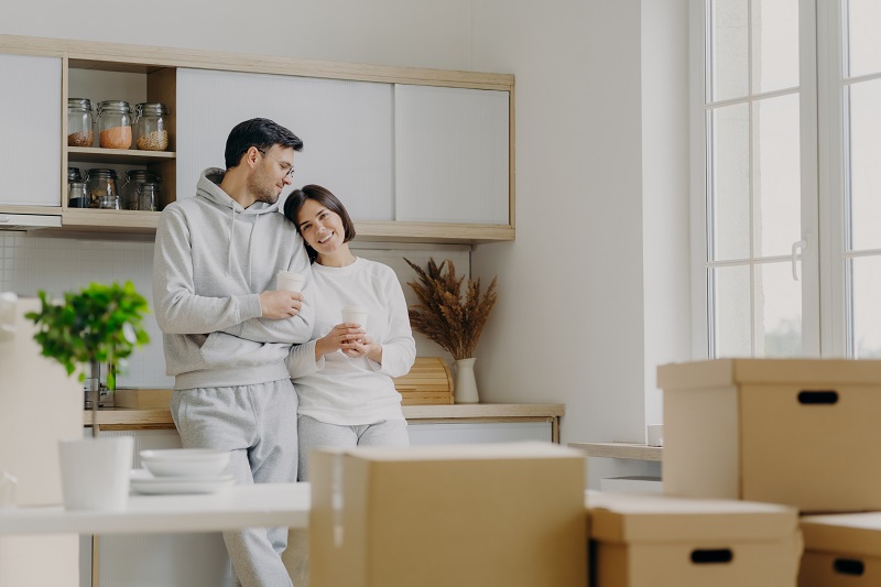 Photo of delighted relaxed husband and wife pose near modern kitchen furniture, have glad expressions, drink takeaway coffee, surrounded with cardboard boxes during relocation day. Mortgage.